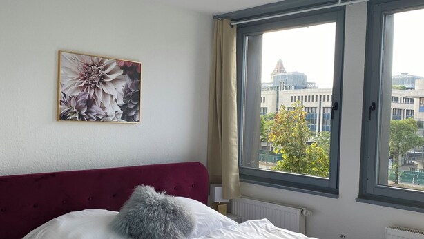A hotel room with a bed, a window with a view of buildings, a ceiling light, and a wall-mounted flower painting. - Modern & central in the cityI 8 guests I kitchen I washing machine I balcony (Stuttgart)