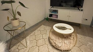 Ein Wohnzimmer mit Fernseher auf einem Schrank, einer Topfpflanze, einem runden Tisch und einem gemusterten Teppich.