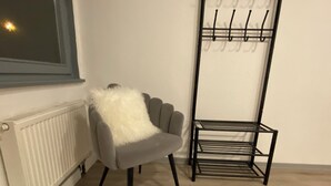 Ein Zimmer mit grauem Sessel, weichem, weißen Überwurf, Heizkörper und einer schwarzen Metallregaleinheit mit Haken.