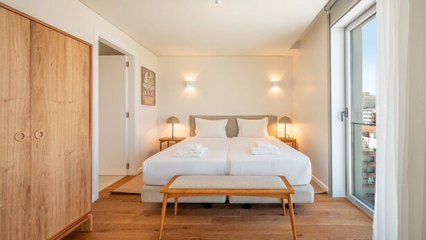 A modern bedroom with a large bed, bedside lamps, a wooden wardrobe, and a city view through a sliding door. - GuestReady - Republic Apartments 8D (Lisbon)