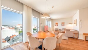 A modern living room with a wooden dining table, comfortable seating, and large windows offering a city view. - GuestReady - Republic Apartments 8D (Lisbon)