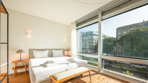 A modern bedroom with a large bed, bedside tables, a bench, and a large window offering a city view. - GuestReady - Republic Apartments 4E (Lisbon)