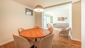 A modern living room with a round wooden dining table, beige chairs, a grey sofa, and a large window. - GuestReady - Republic Apartments 8E (Lisbon)