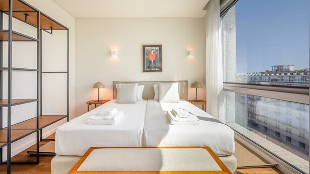 A hotel room with a large bed, two bedside tables, a small sofa, a wooden bench, and a large window offering a city view. - GuestReady - Republic Apartments 8E (Lisbon)