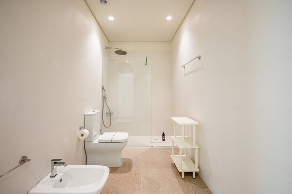 A modern bathroom with a toilet, sink, shower, and a small shelf unit.