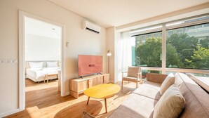A modern hotel room with a large window, a flat-screen TV, a wooden coffee table, a sofa, and a bed with white bedding.