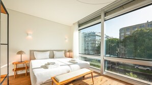 A modern bedroom with a large bed, bedside tables, a bench, and a large window offering a city view.