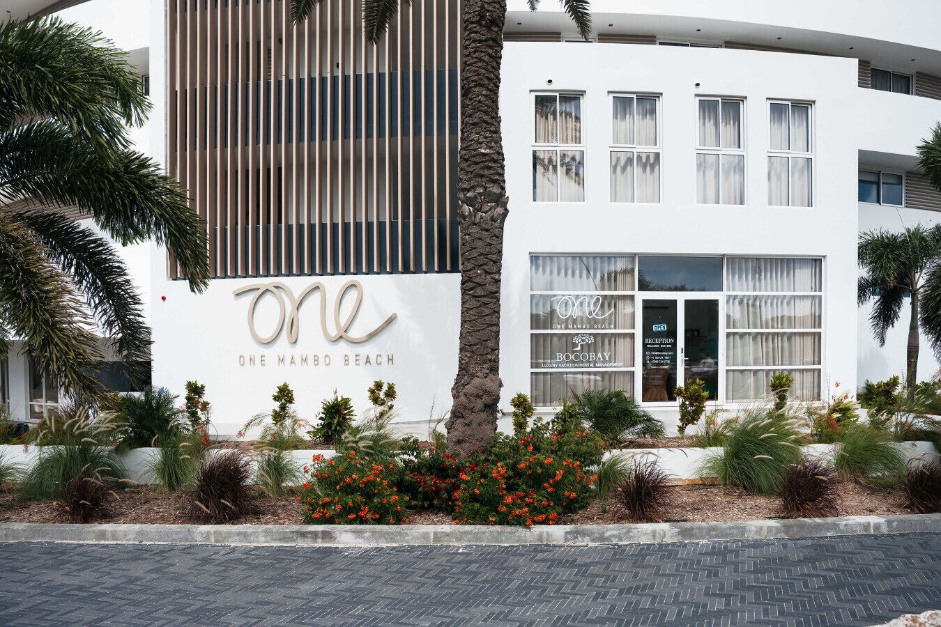 Un bâtiment moderne avec une enseigne indiquant « One Mambo Beach » et « BOCOBAY, gestion de locations de vacances de luxe ».