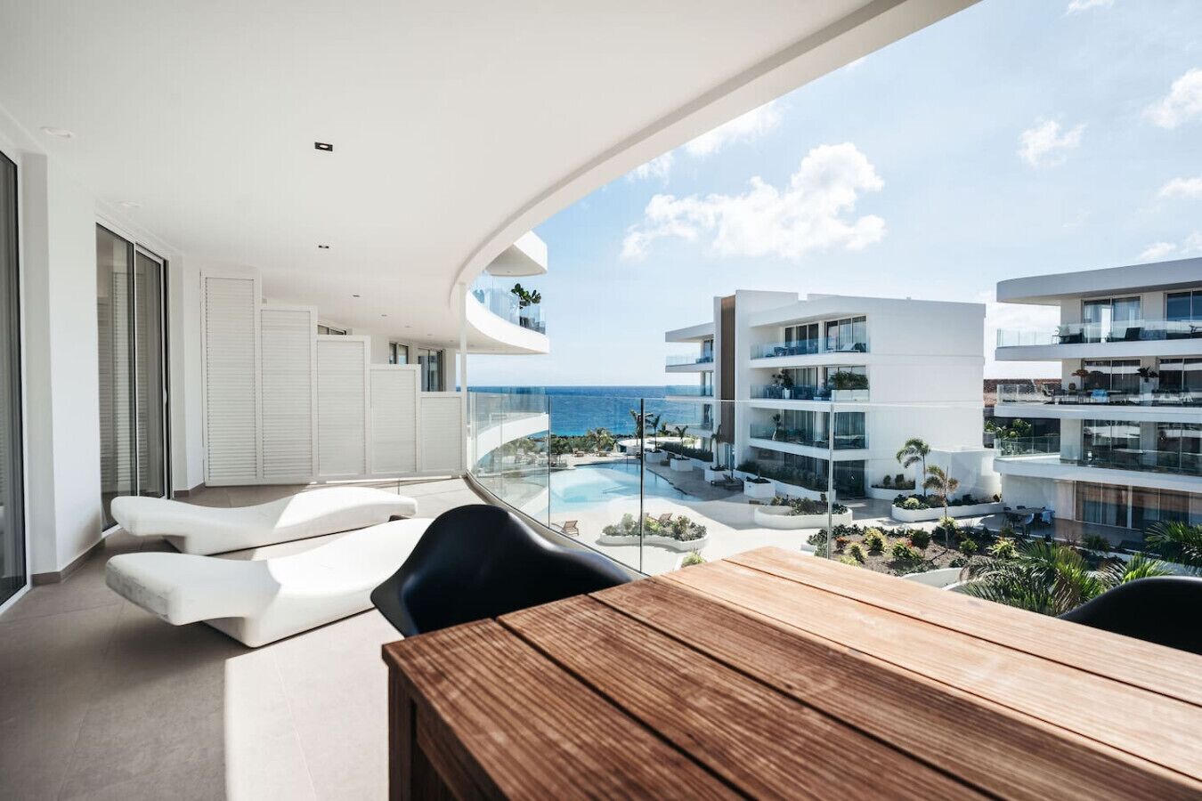 Een modern balkon met een houten tafel en witte loungestoelen, met uitzicht op een zwembad en andere gebouwen.