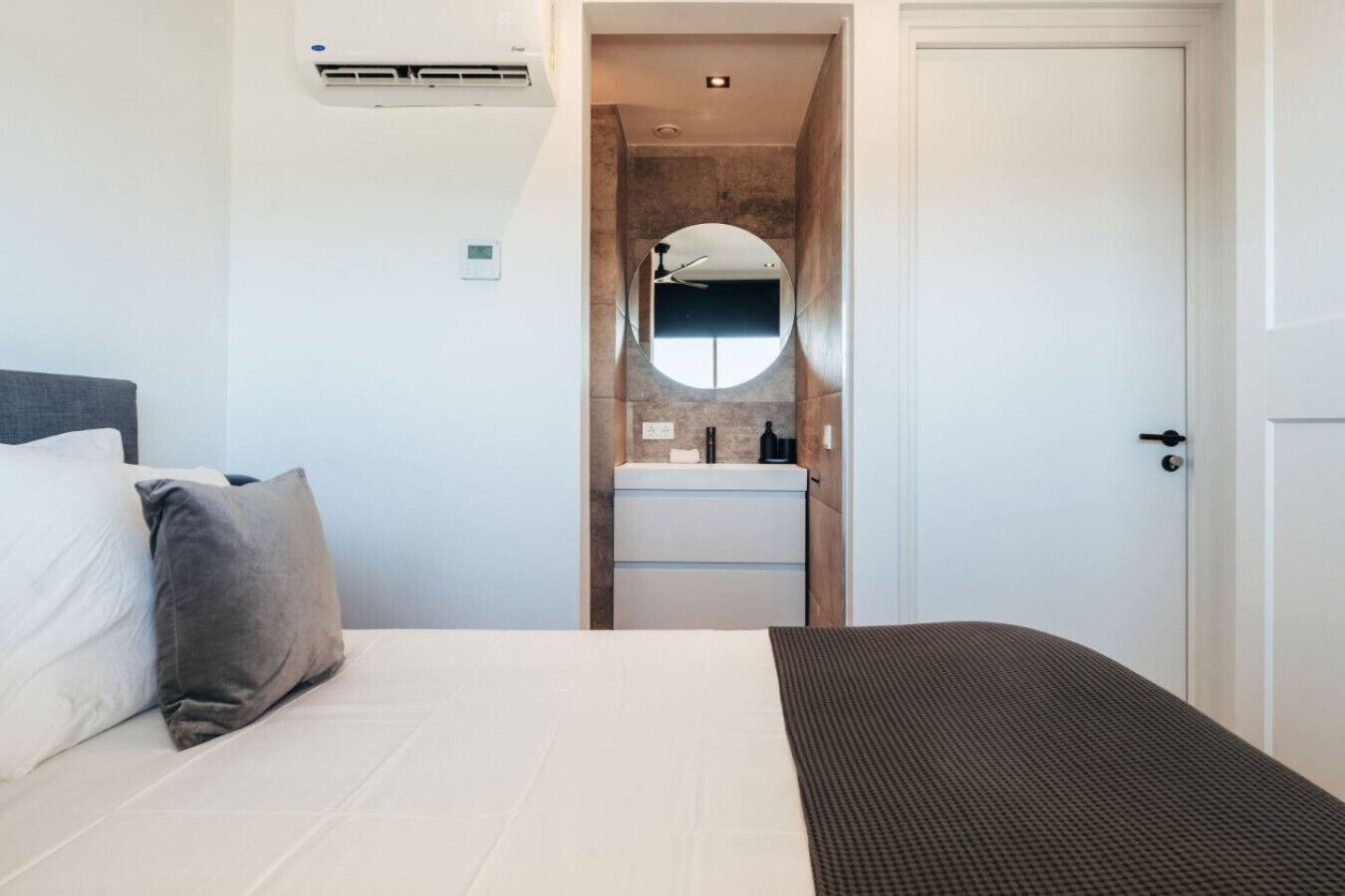 A bedroom with a bed, a bedside table, a mirror, and a bathroom with a sink and mirror.