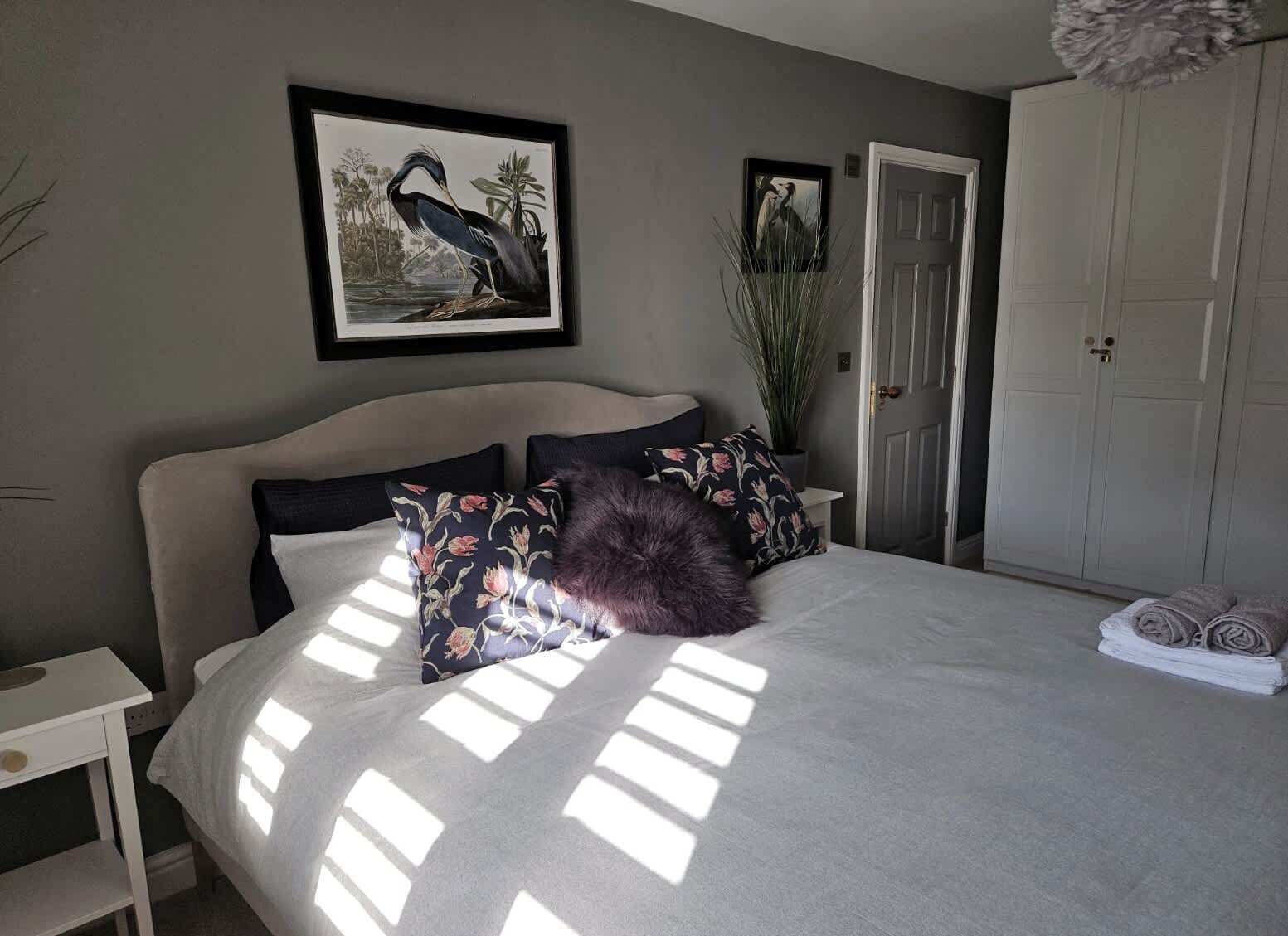 Un dormitorio con cama, almohadas, una mesita de noche, una puerta y una planta.