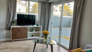 A modern living room with a glass coffee table, a sofa with colorful cushions, a TV on a white cabinet, and a view of a courtyard with yellow flowers.
