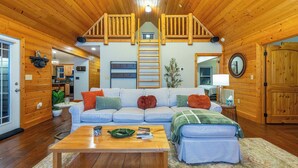 Ein gemütliches Blockhaus-Interieur mit Holzbalkendecke, einer zentralen Wohnbereits mit Sofa und Couchtisch sowie einer sichtbaren Küche im Hintergrund.