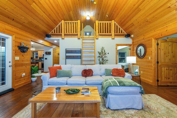 L’intérieur chaleureux d’un chalet avec un plafond en bois, un espace de vie central comprenant un canapé et une table basse, et une cuisine visible au fond.