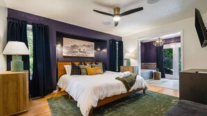 A bedroom with a large bed, wooden headboard, and a ceiling fan. There is a window with curtains, a nightstand with a lamp, and a painting on the wall.