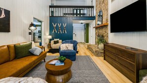 A spacious living room with a high ceiling, a wooden coffee table, a sofa, a blue armchair, and a TV mounted on the wall.