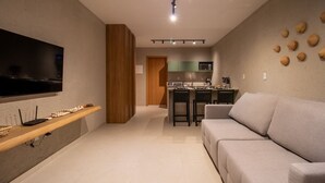 Uma sala de estar moderna com uma TV de tela plana, uma estante de madeira, um sofá cinza e uma cozinha ao fundo.