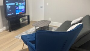 A living room with a blue armchair, a grey sofa, a small coffee table, and a TV on a stand. - Elegant 2-Bed 2-Bath SE Calgary, fully furnished near amenities, mid-term, ready (Calgary)