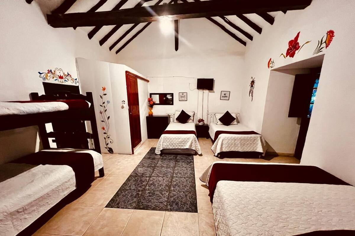 Ein Hotelzimmer mit vier Betten, die nebeneinander liegen, einer kleinen Sitzecke mit Tisch und Stühlen sowie einem an der Wand befestigten Fernseher.