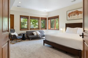Una camera da letto con un letto grande, un divano, una sedia e un tavolino. Quattro finestre offrono luce naturale e vista sugli alberi.