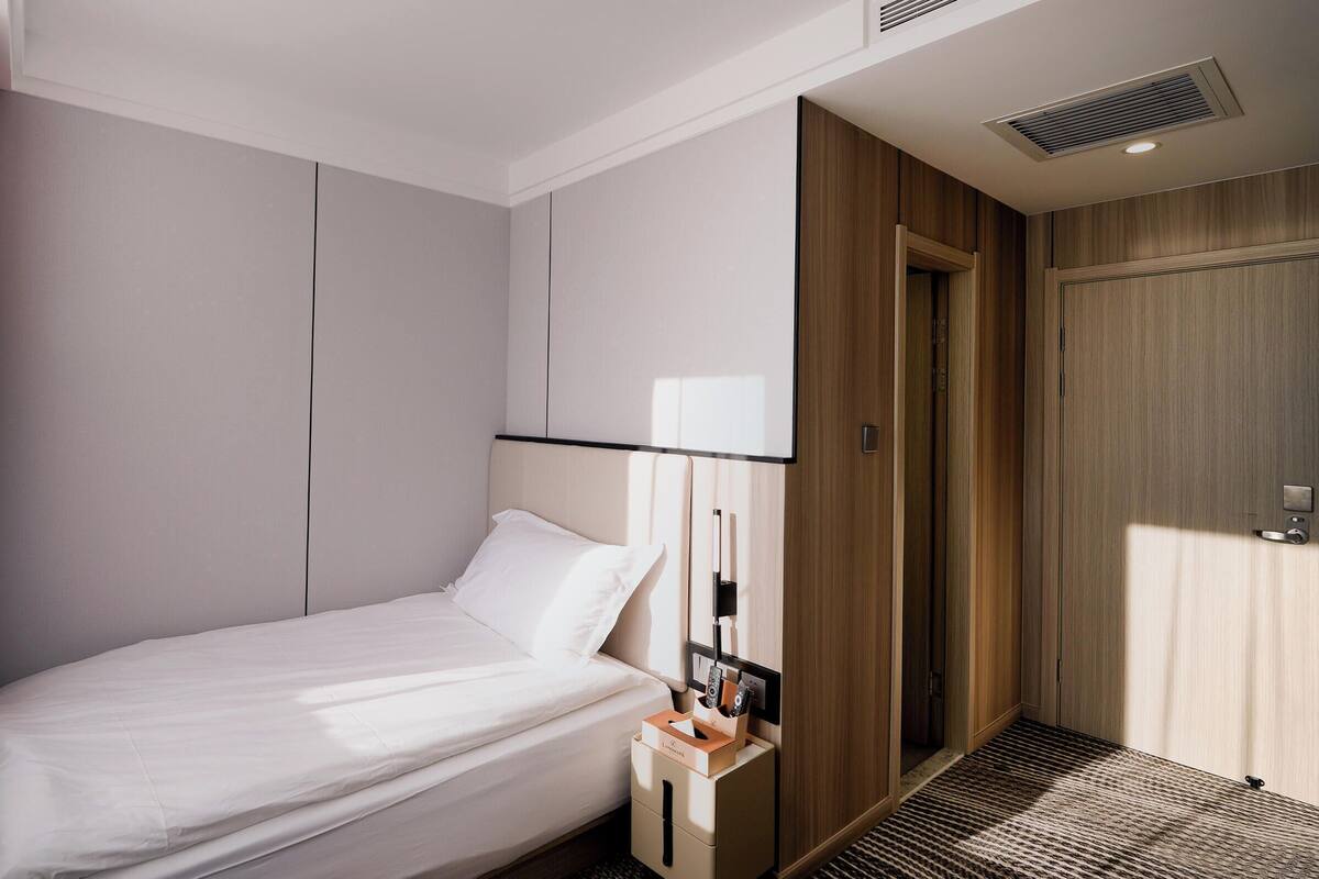 Habitación de hotel moderna con una cama bien hecha, una mesita de noche con lámpara, un armario y una puerta que da a otra habitación.