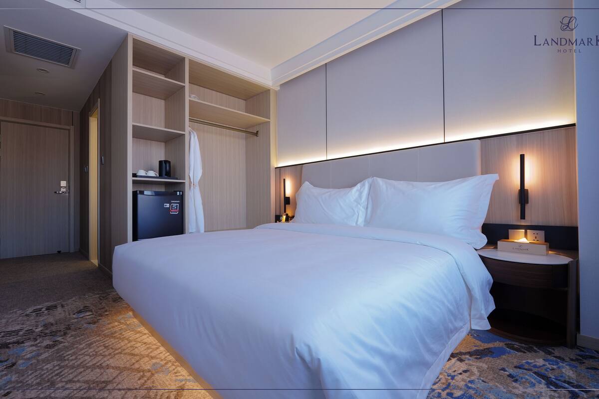 Habitación de hotel con una cama grande, un minibar, un armario y una mesita de noche con lámpara.