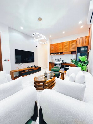 Una sala de estar moderna con una mesa de centro de vidrio, sofás blancos, un televisor de pantalla plana, armarios de madera y una zona de cocina con microondas y refrigerador.