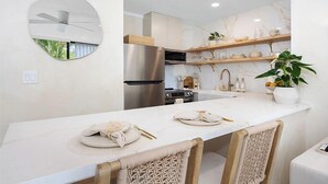 A modern kitchen with a central island, built-in appliances, and a hanging shelf with decorative items. - Kaia Villa, at Turtle Bay - Modern North Shore Oahu Retreat w/Lanai & Golf Views (Kahuku)