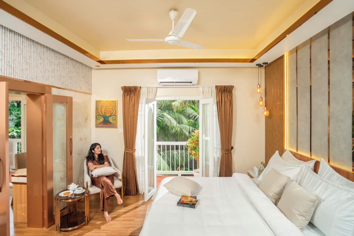 A hotel room with a bed, a sitting area with a table, and a balcony with a view of greenery.