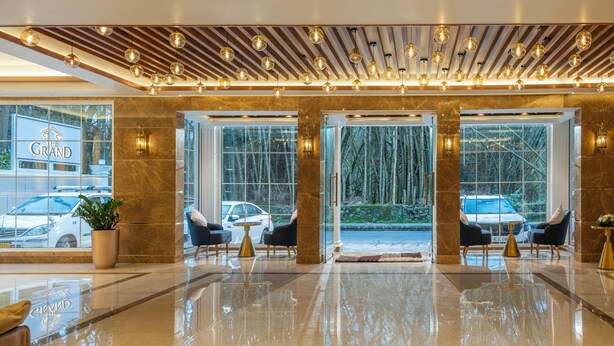 A modern hotel lobby with a polished marble floor, two seating areas with armchairs, and a ceiling with hanging spherical light fixtures. - The Grand (Thekkady)