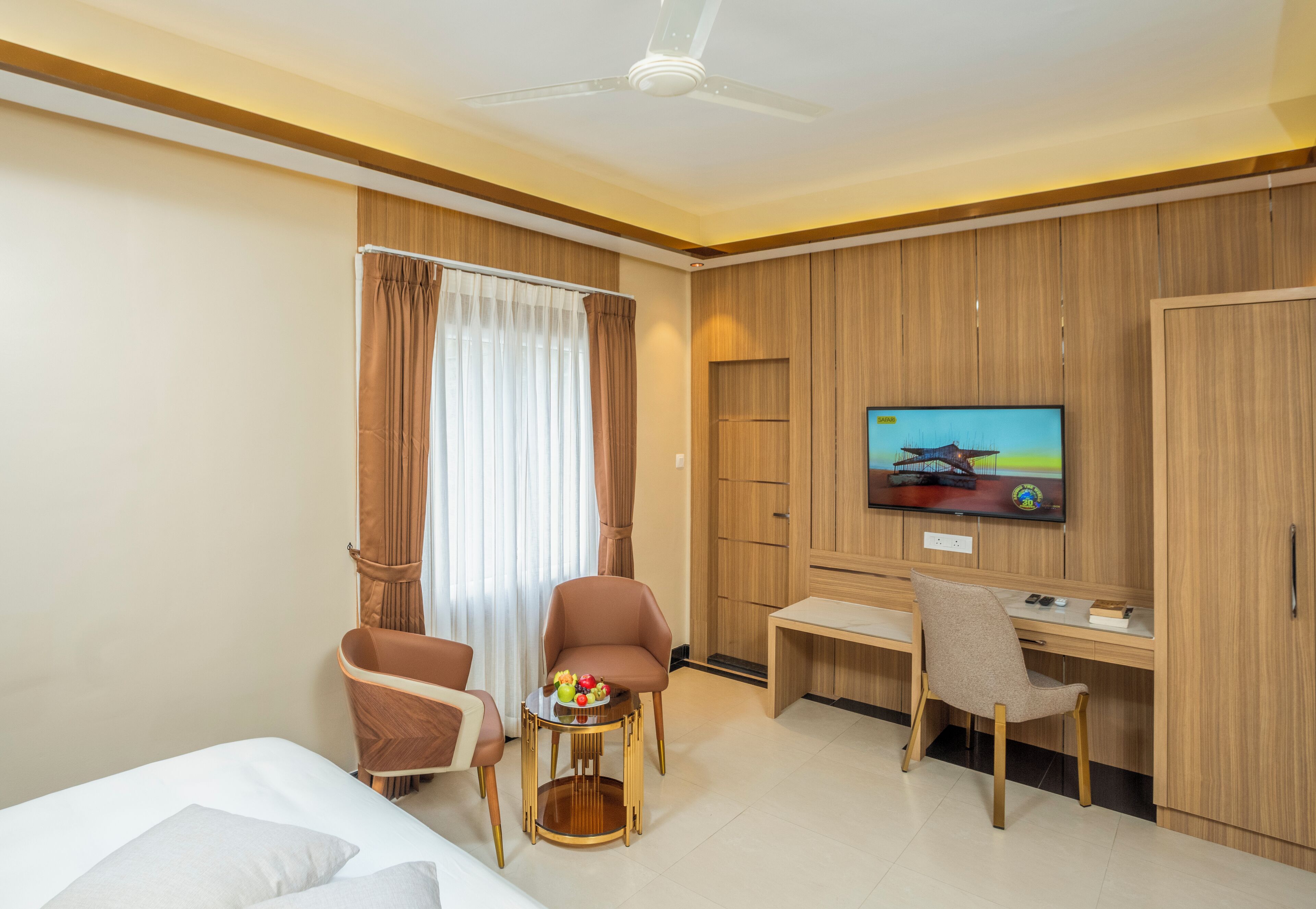 A hotel room with a bed, two armchairs, a small table with fruit, a desk with a chair, a television, and wooden paneling on the walls.