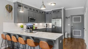 Een moderne keuken met grijze kastjes, roestvrijstalen apparatuur, een marmeren eiland met barkrukken, hangende verlichting en een decoratieve wandversiering.