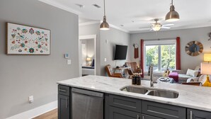 Een moderne keuken met een marmeren aanrechtblad, donkere kasten en een inbouw vaatwasser. Er is een eethoek met een tafel en stoelen, en een groot raam met uitzicht op groen.