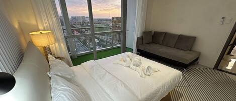 A hotel room with a bed, a sofa, a window with a city view, and a lamp.