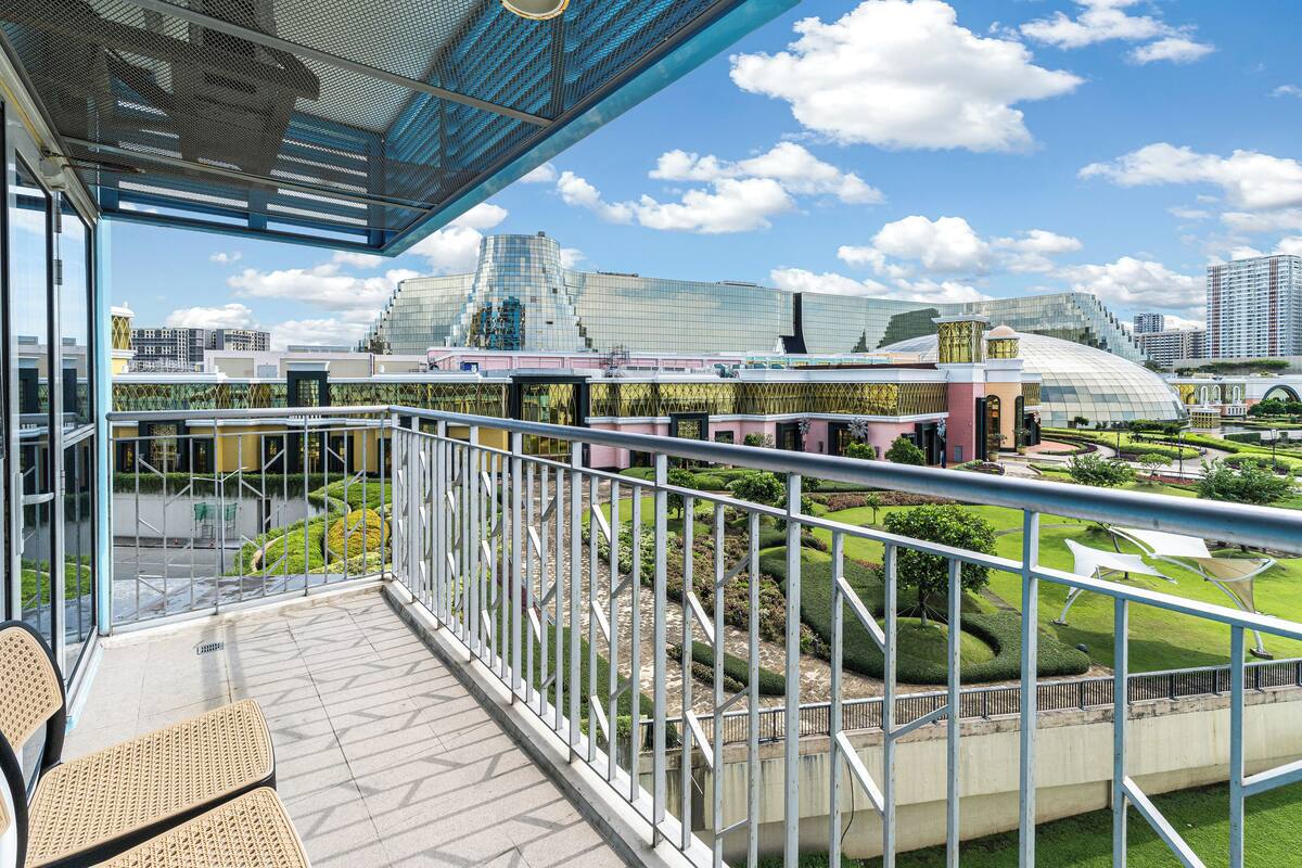 Ein Balkon mit Korbstühlen, einer Glastrennwand und Blick auf einen modernen Gebäudekomplex mit Glaskuppel und Grünflächen.