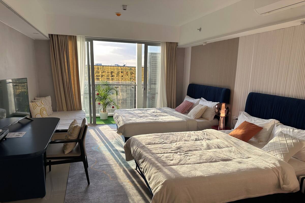 Ein Hotelzimmer mit zwei Betten, einem Schreibtisch, einem Stuhl und einem großen Fenster mit Blick auf Gebäude.