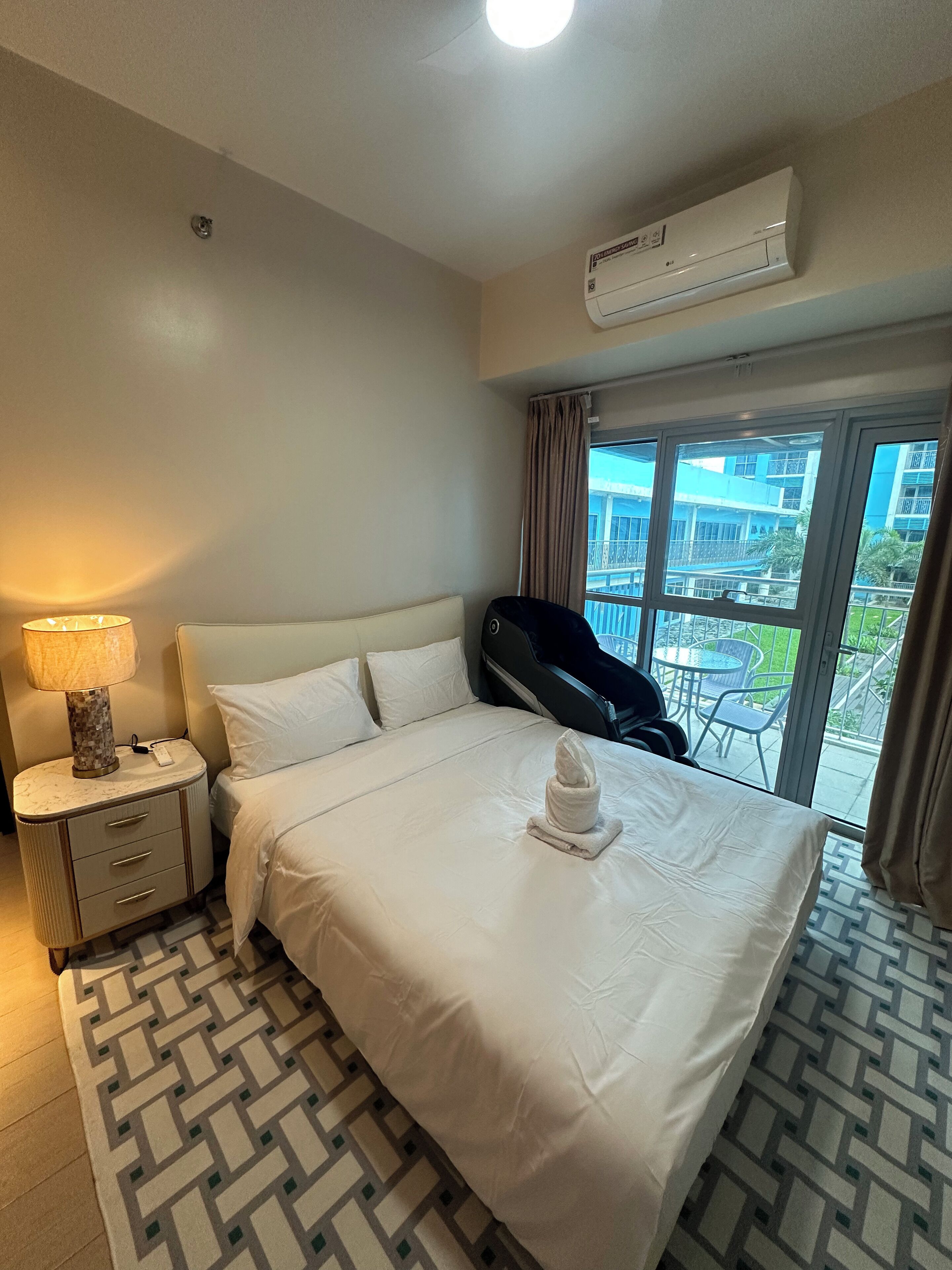 A hotel room with a bed, a bedside lamp, a window with a view of buildings, and an air conditioning unit.