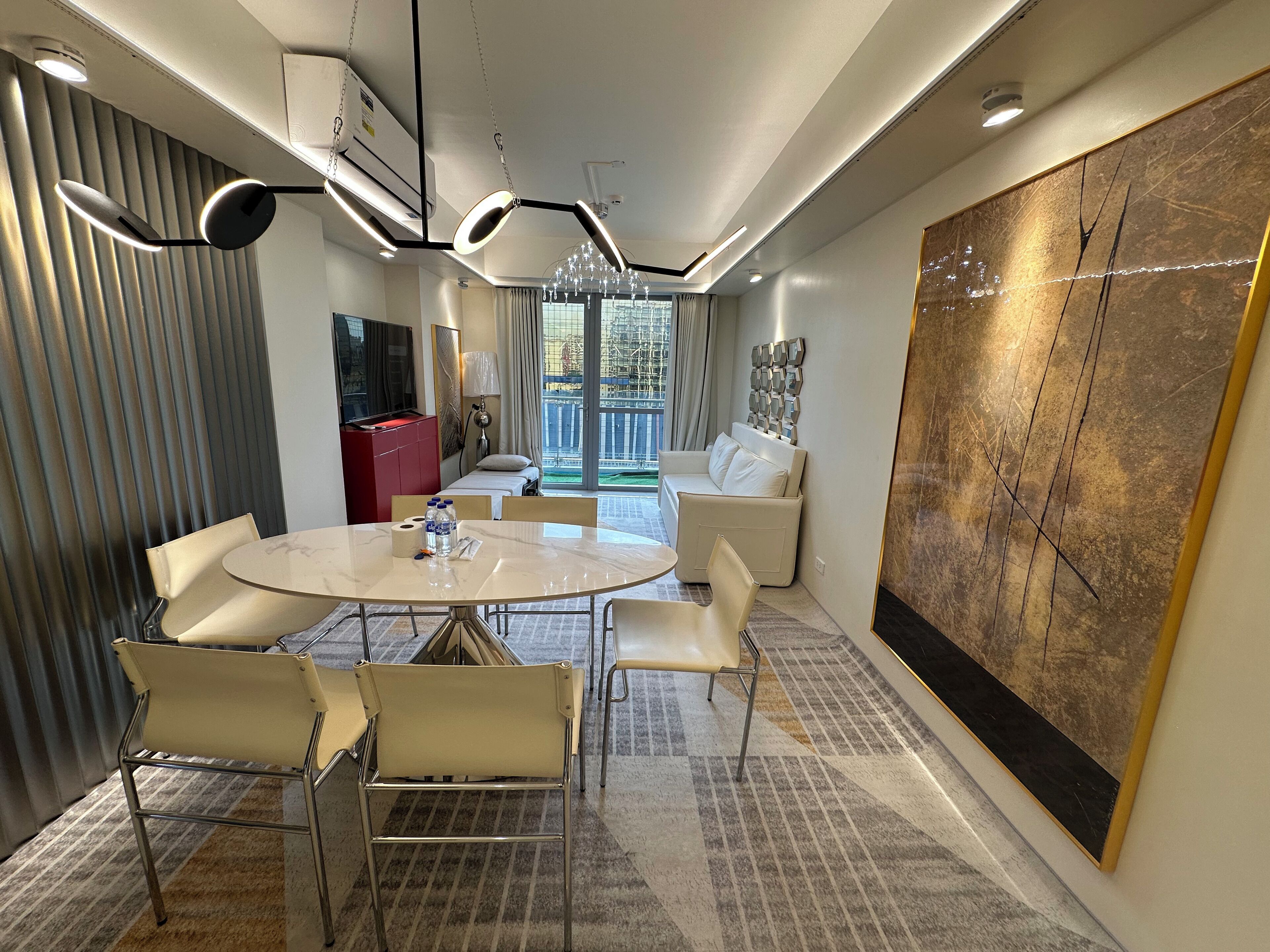 A modern hotel room with a round dining table, white chairs, a red minibar, a large abstract painting, and a view of the city through large windows.