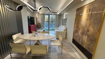 A modern hotel room with a round dining table, white chairs, a red minibar, a large abstract painting, and a view of the city through large windows.