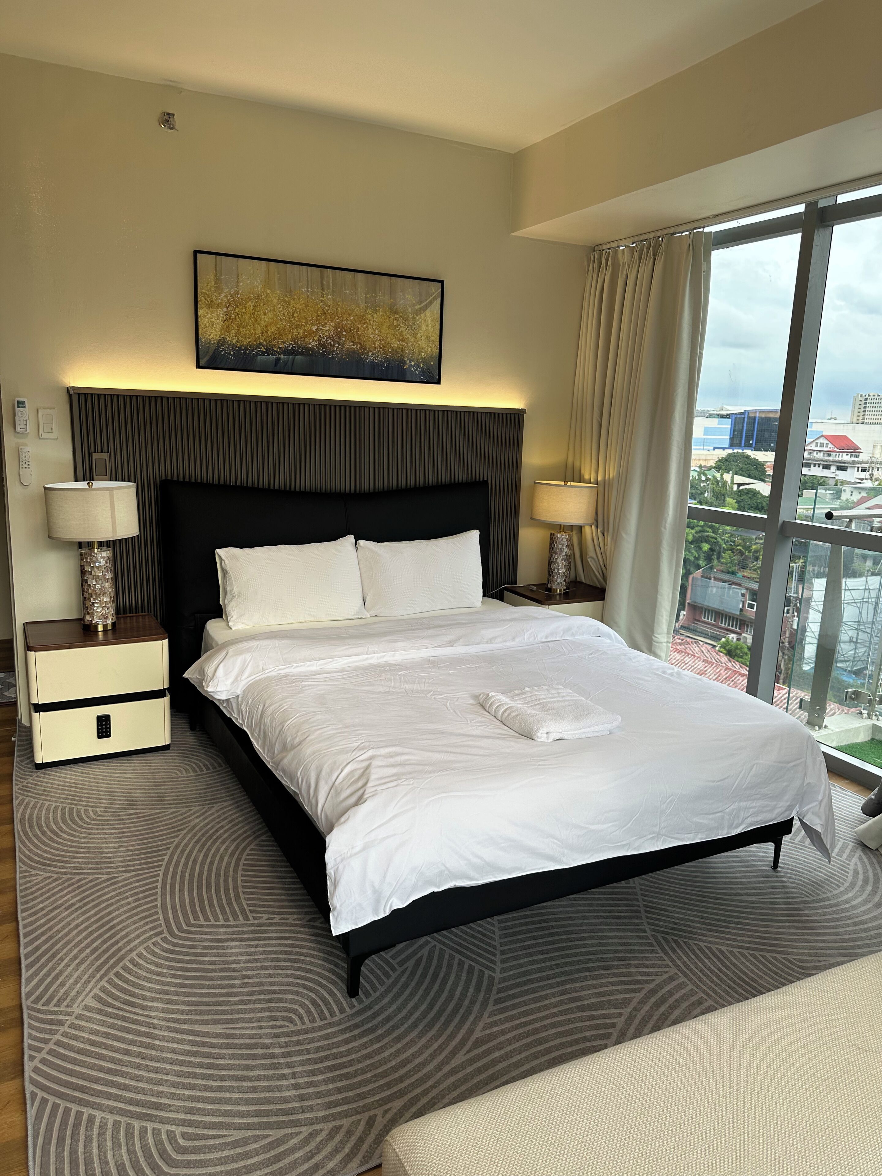 A hotel room with a large bed, two bedside tables, a headboard, a painting, and a view of buildings through the window.