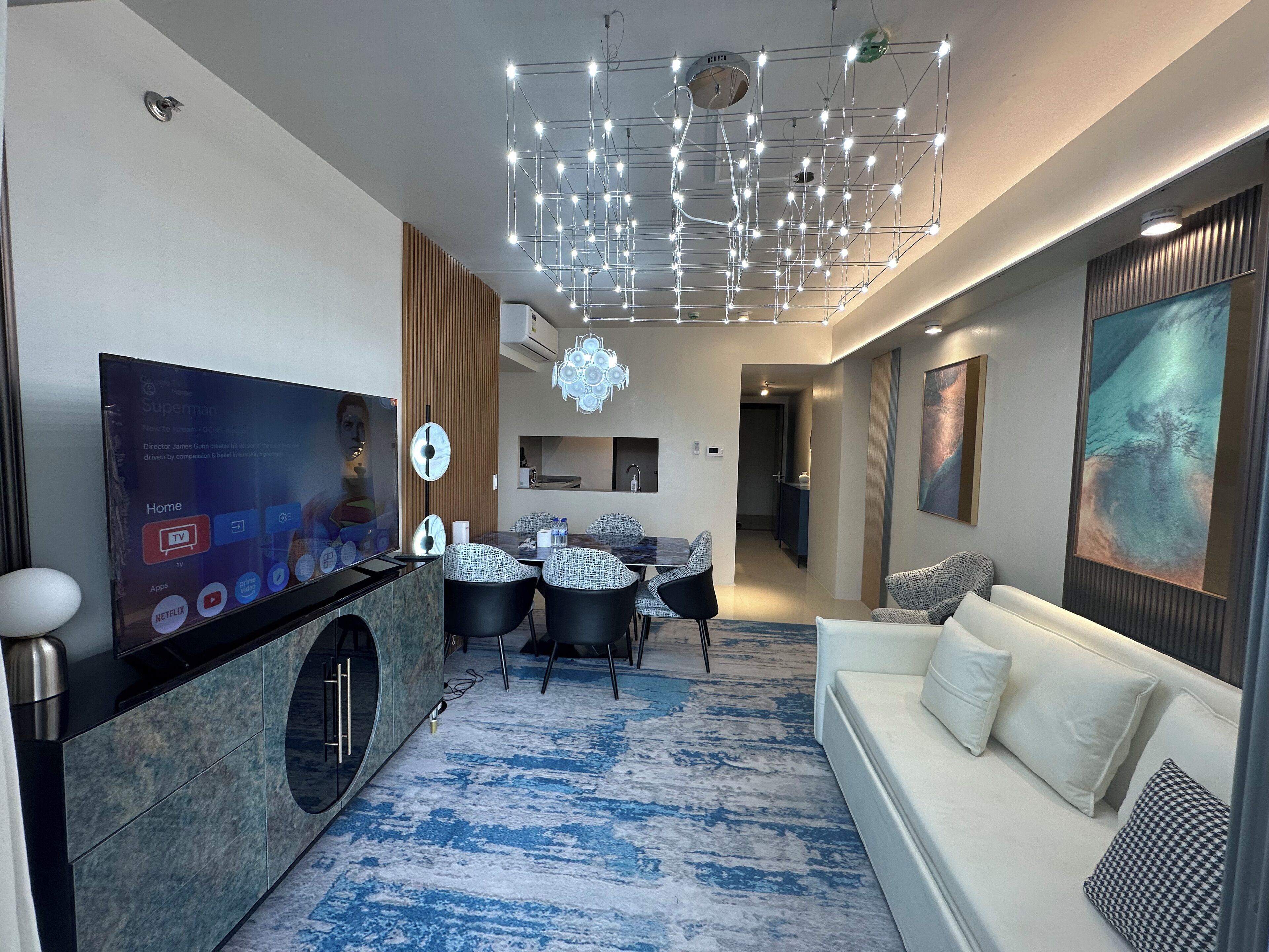 A modern living room with a flat-screen TV, a dining area with a table and chairs, a white sofa, and a patterned carpet.