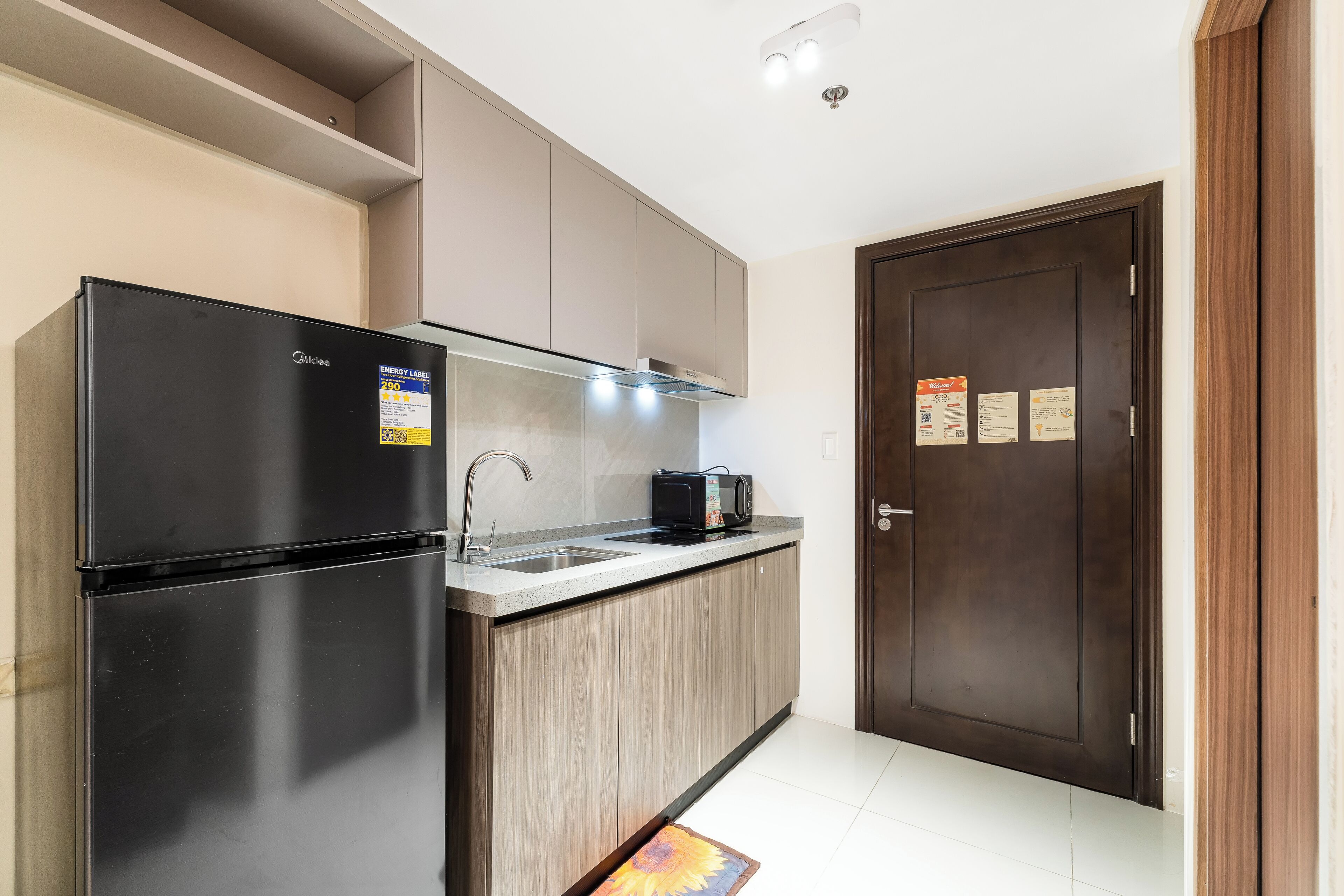 A modern kitchen with a black refrigerator, a microwave, and a sink. There is a wooden cabinet above the counter and a dark wooden door to the right.