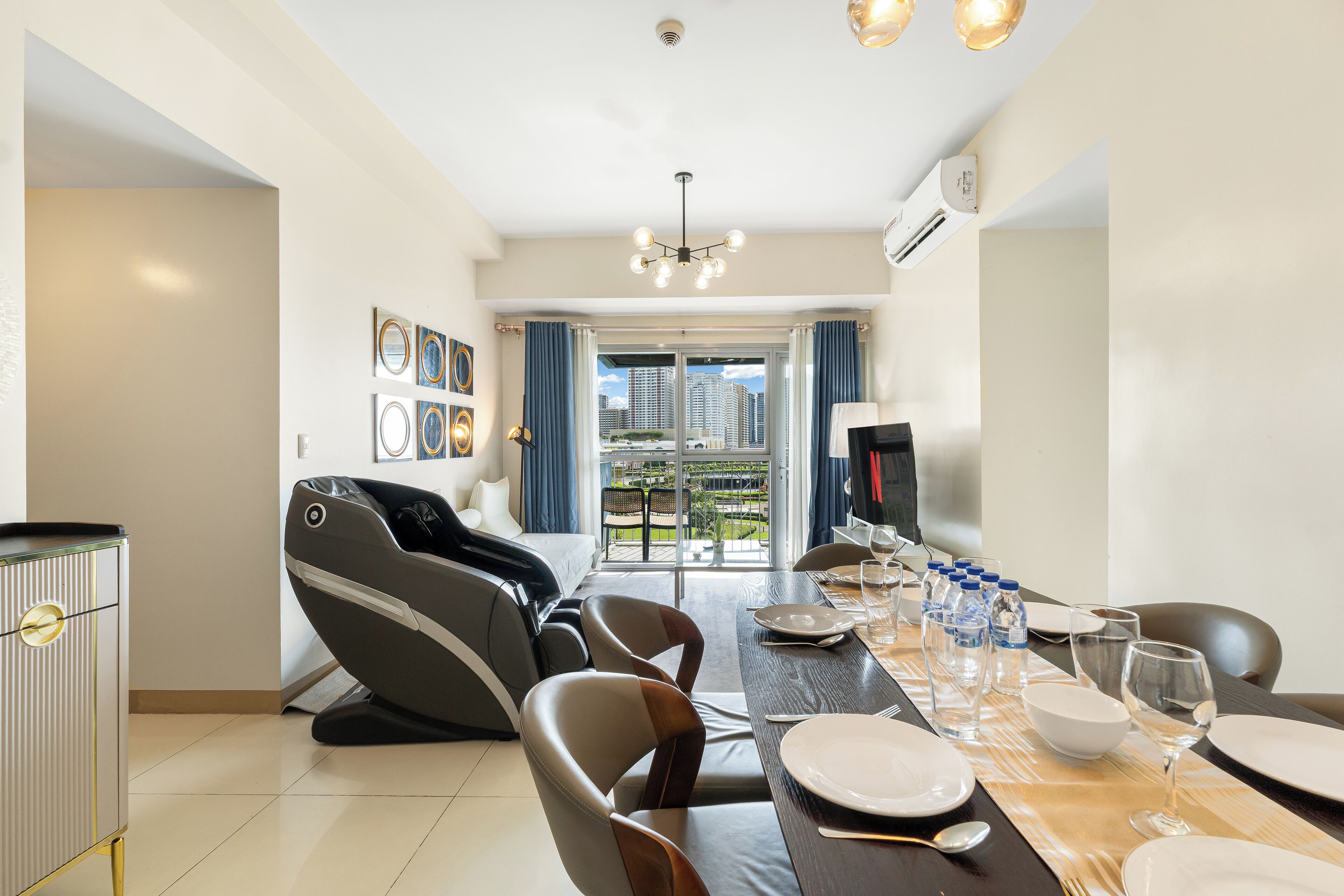 A modern living room with a massage chair, dining table set for four, and a large window with a city view.