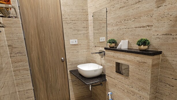 A bathroom with a wooden door, a white sink, a mirror, and a small shelf with plants. - Hotel Siyaram (Ayodhya)