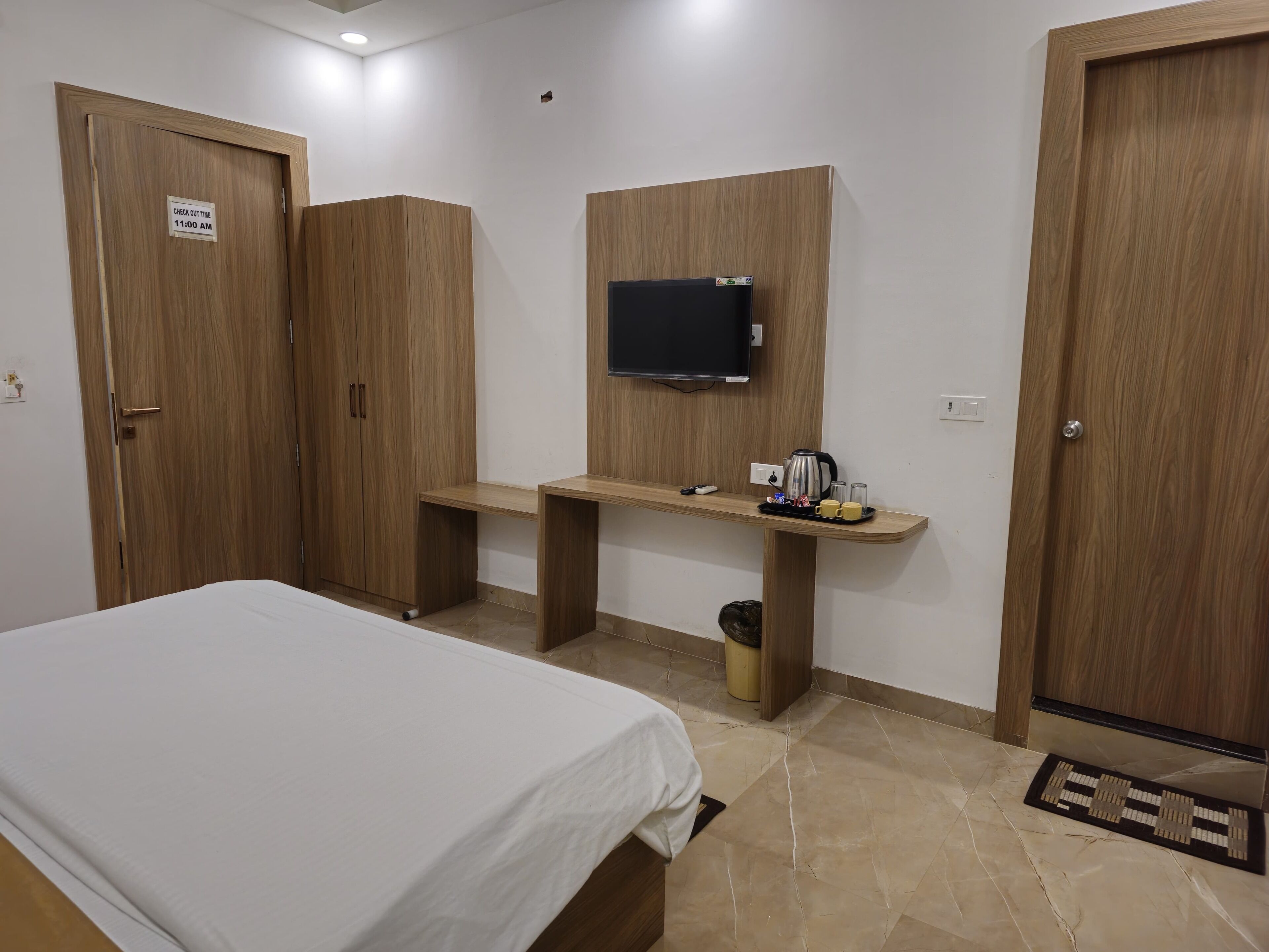 A hotel room with a bed, a desk with a flat-screen TV, wooden wardrobes, and a check-out sign.