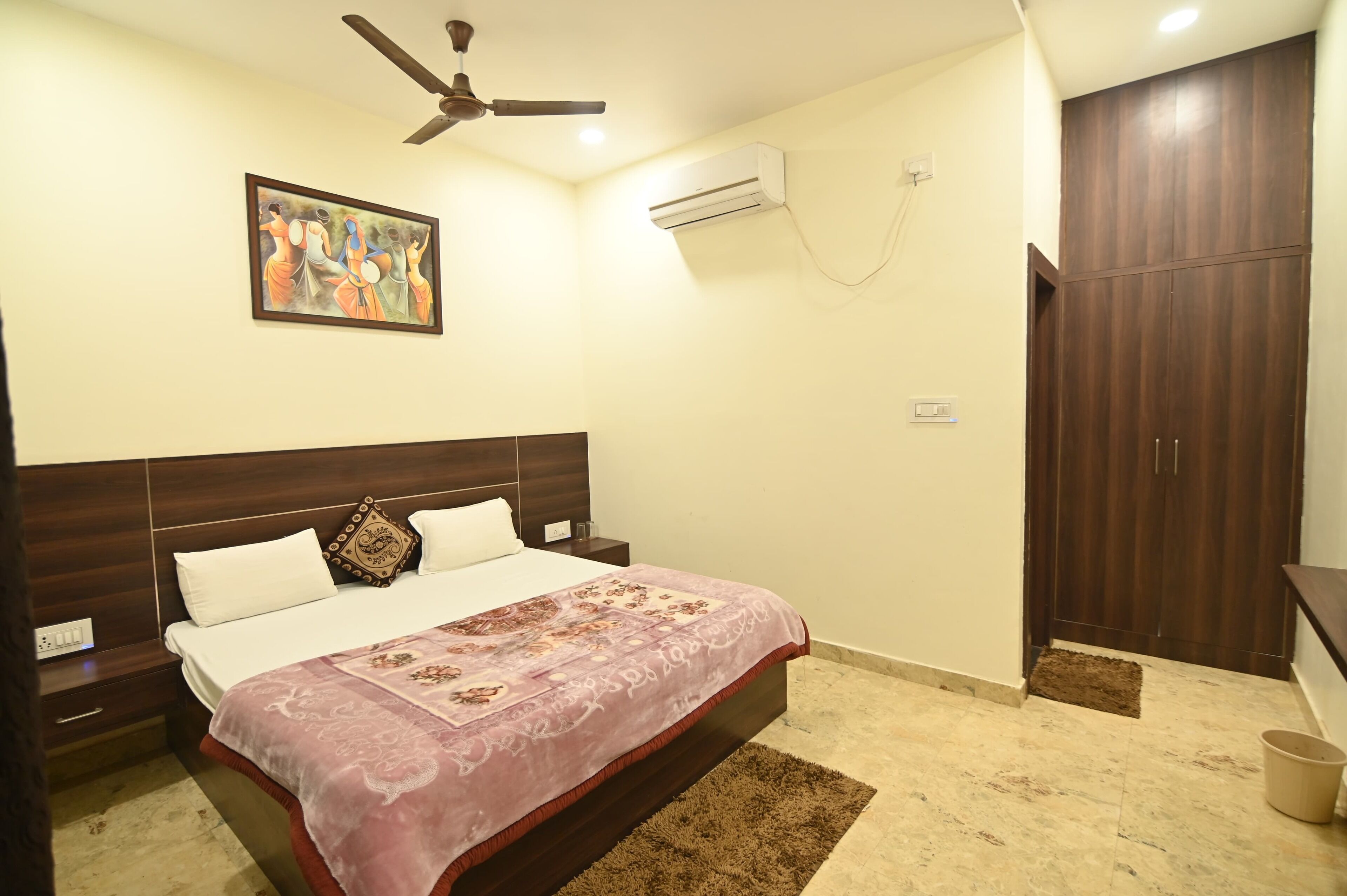 A bedroom with a bed, a ceiling fan, an air conditioner, a wardrobe, and a painting on the wall.