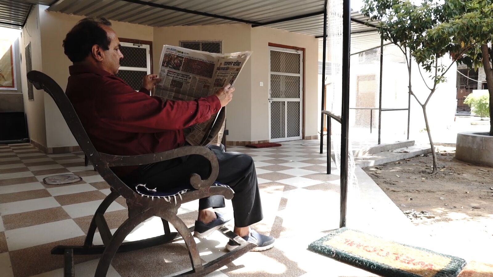 Iemand zit op een metalen bankje en leest een krant in een overdekte buitenruimte met een betegelde vloer en een kleine boom in de buurt.