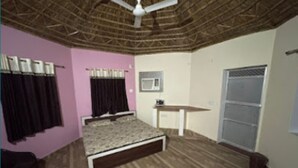 A bedroom with a bed, a ceiling fan, a small table, a window with a metal screen, and a door. - Shyam Luxurious Huts & Guest House (Shri Madhopur)