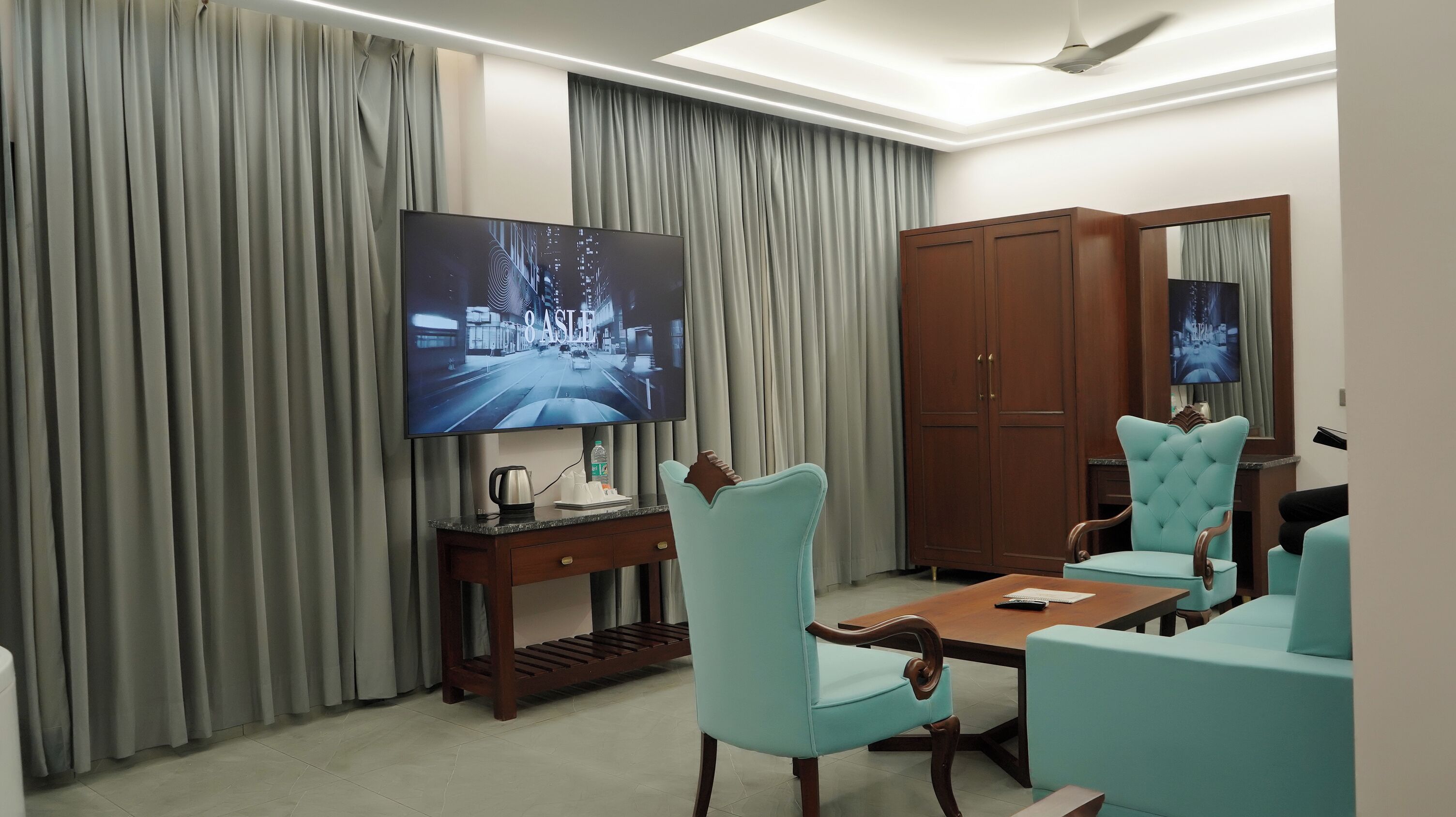 A hotel room with a flat-screen TV, a wooden wardrobe, a dining table with chairs, and a ceiling fan.