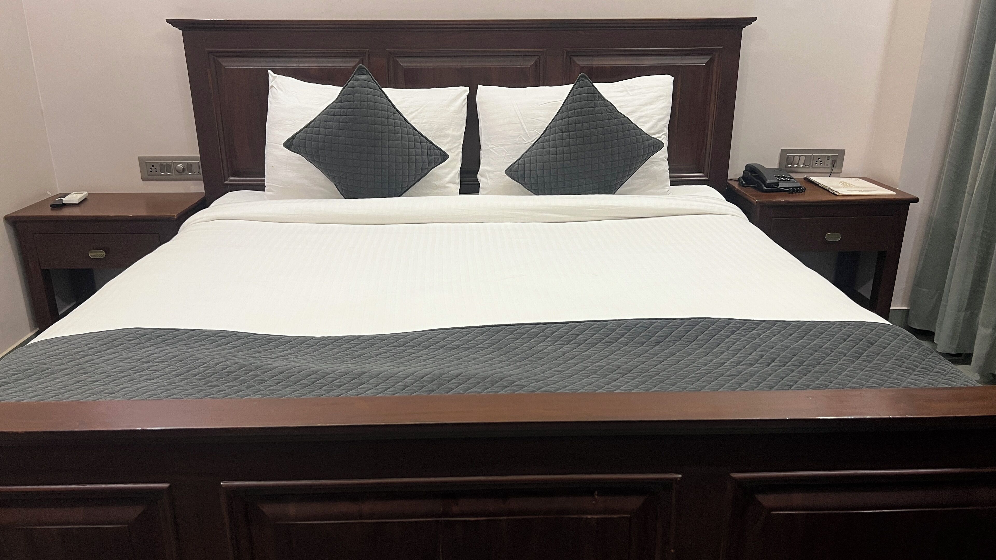Een netjes opgemaakt bed met een donker hoofdbord, twee nachtkastjes met een telefoon en notitieblok, en een stopcontact aan de muur.